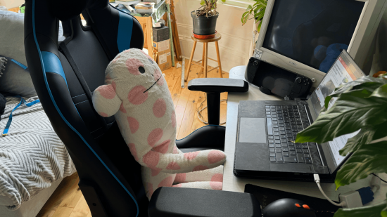 A plushie sat on a gaming chair at a desk with laptop, CRT monitor and plants